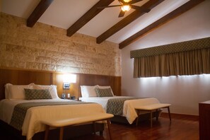Una habitación con dos camas, vigas de madera en el techo, pared de piedra, ventana con cortinas y una mesita de noche con lámpara.