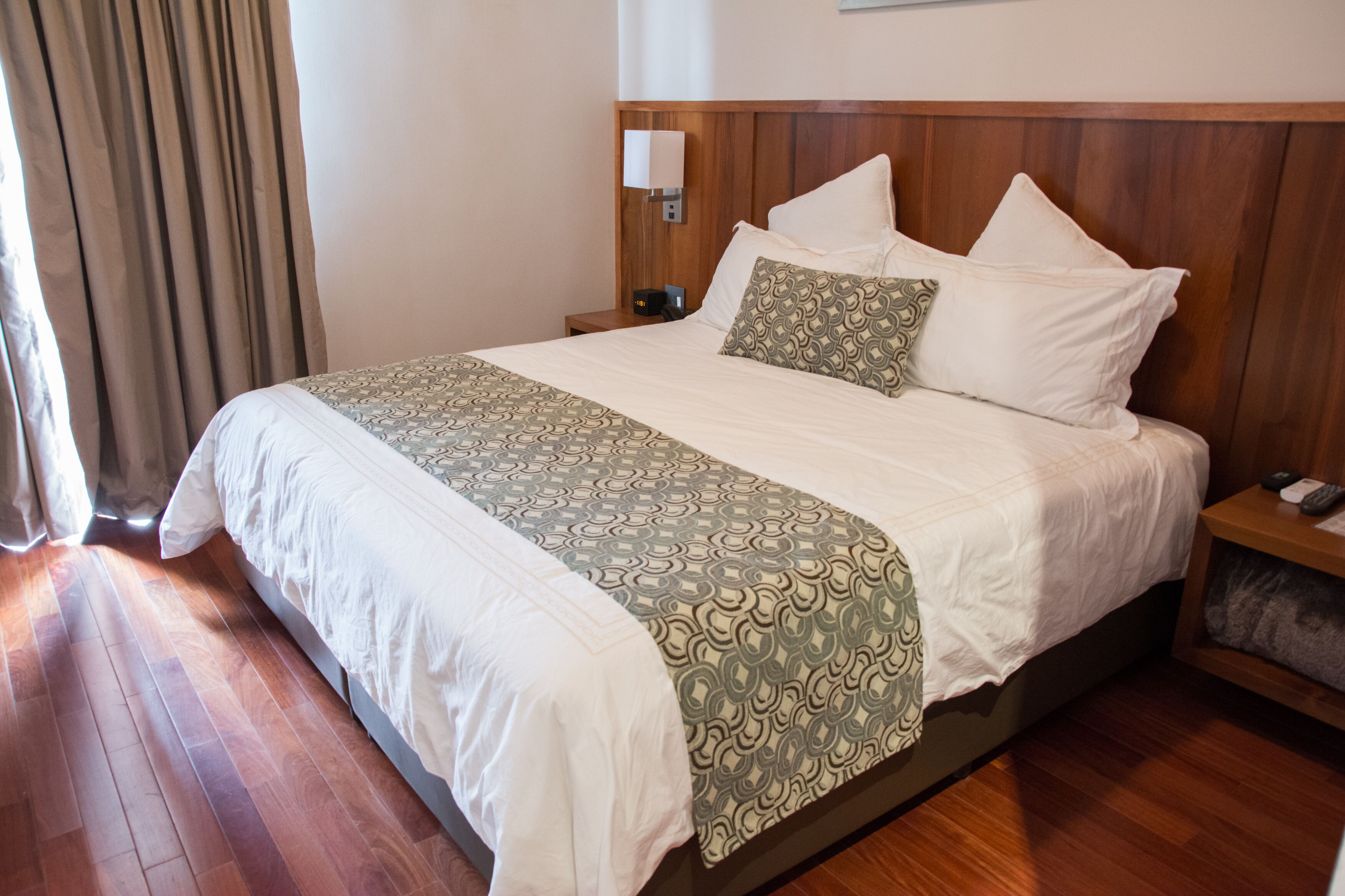 A neatly made bed with a patterned bedspread, a bedside table with a lamp, and a nightstand with a remote control.
