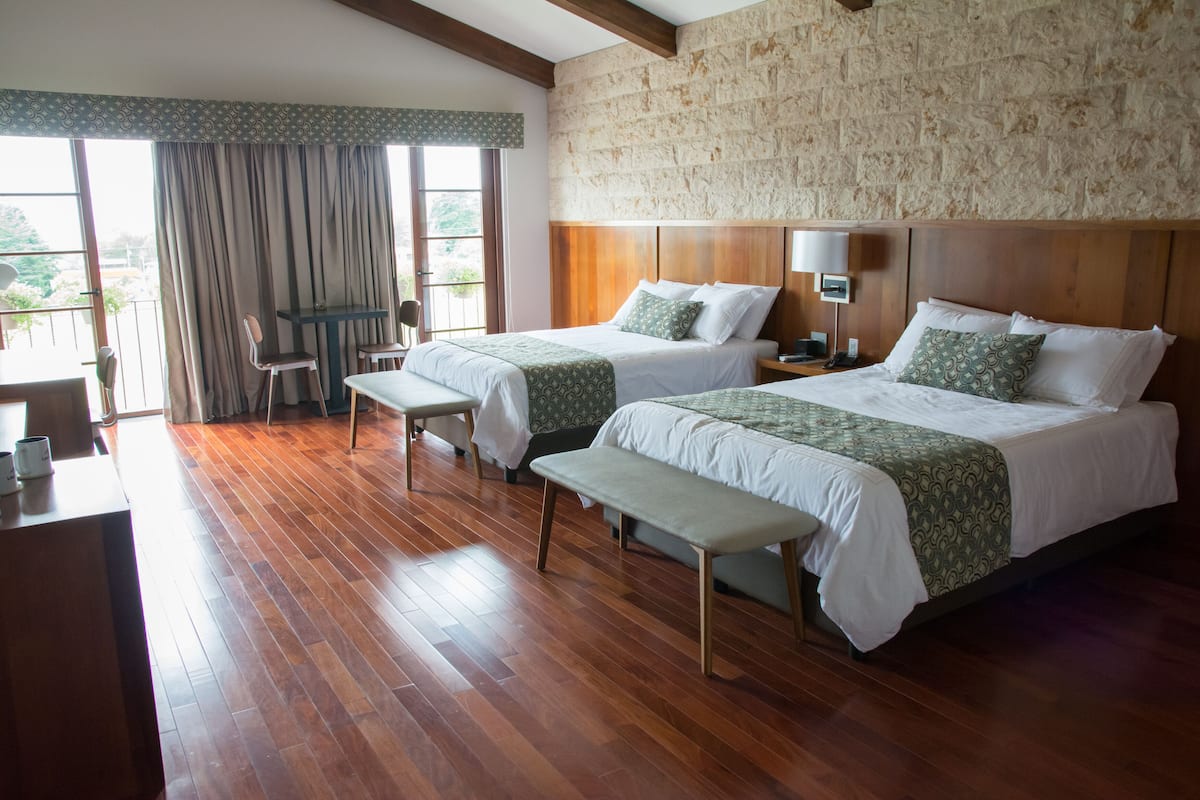 A hotel room with two beds, wooden flooring, a balcony with curtains, and a stone wall.