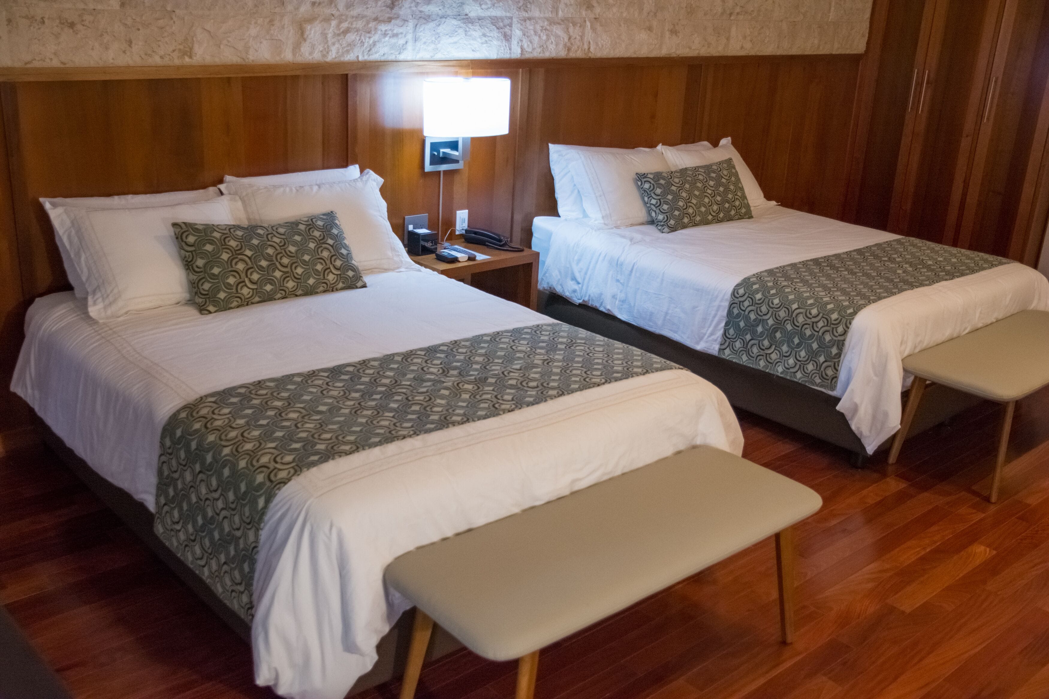 Une chambre d’hôtel avec deux lits, chacun agrémenté d’une couvre-lit et de coussins à motifs, une table de chevet avec une lampe et un banc au pied des lits.