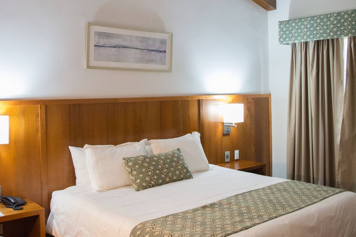 A hotel room with a wooden ceiling, a bed with a patterned bedspread, two bedside lamps, and a ceiling fan.