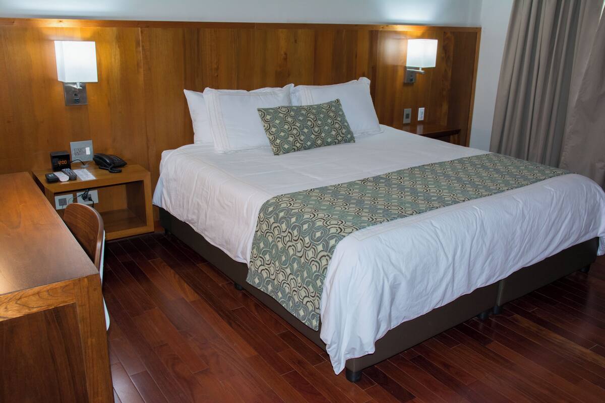 A hotel room with a wooden headboard, a bed with white linens, a patterned throw blanket, a bedside table with a phone and a lamp, and a wooden floor.