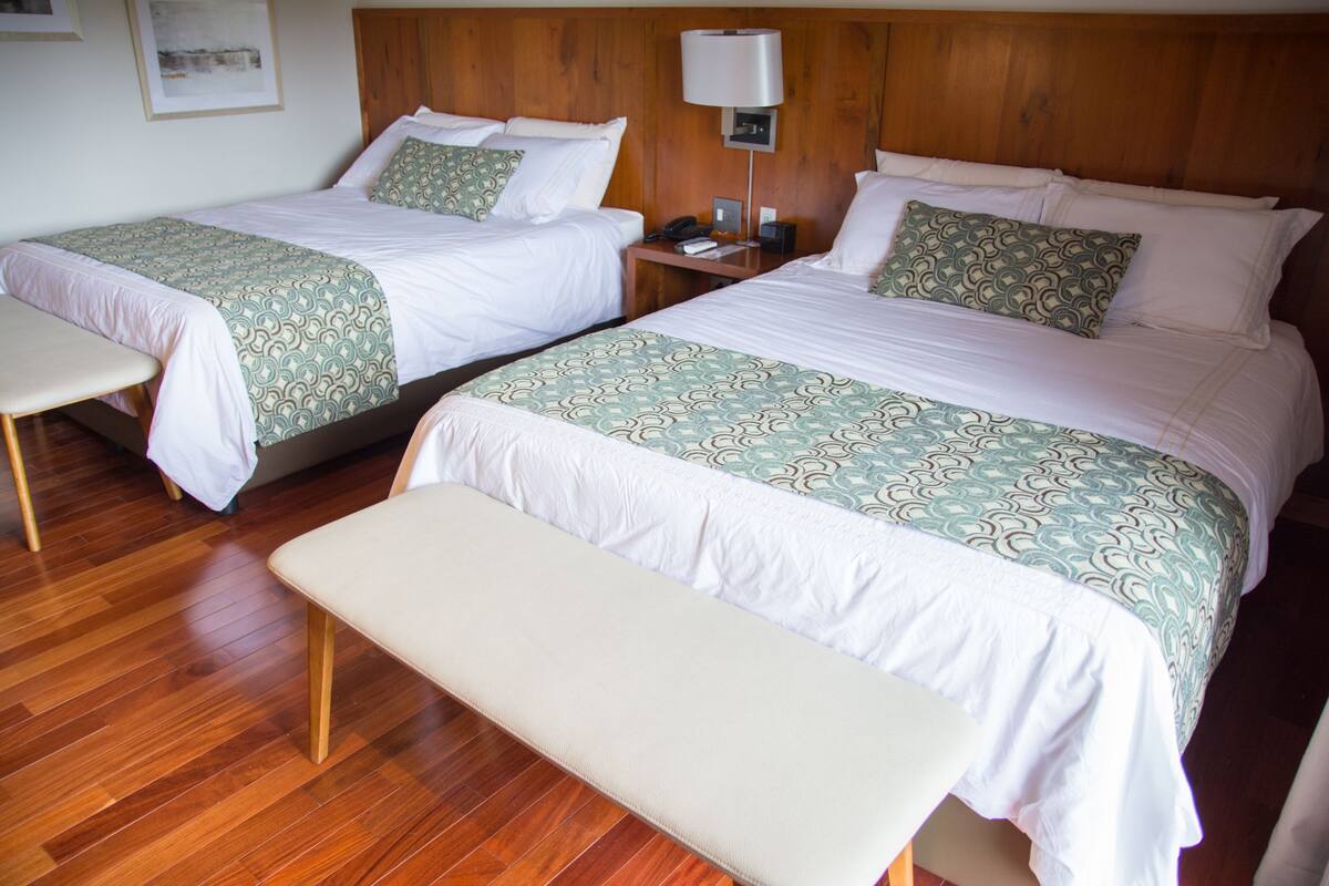 A hotel room with two beds, a bench, wooden flooring, and a nightstand with a lamp.