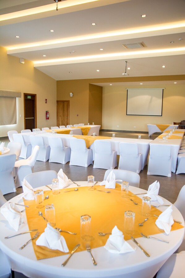Un salón de conferencias con mesas redondas, sillas blancas y una pantalla grande en la pared.