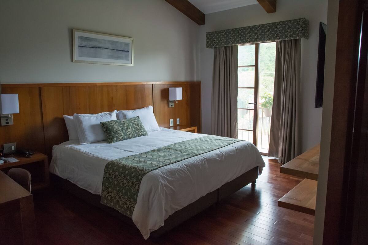 A hotel room with a large bed, wooden headboard, bedside lamps, a desk, and a window with curtains.