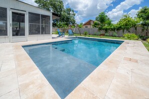 A rectangular swimming pool with a tiled deck, a covered patio area with chairs, and a wooden fence in the background. - Contemporary Oaks Heated Pool + HOT TUB! (Tampa)