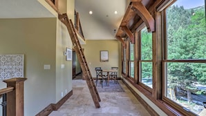 Un couloir avec un escalier en bois, un coin repas avec une table et des chaises, et de grandes fenêtres donnant sur une zone boisée.