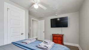 A bedroom with a bed, a ceiling fan, a TV mounted on the wall, and a wooden dresser.