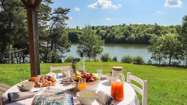 Mesa al aire libre con vista a un lago y bosque, mostrando una comida con frutas, pan y una bebida.