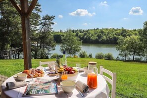 A table set outdoors with a view of a lake and forest, featuring a meal with fruits, bread, and a drink.