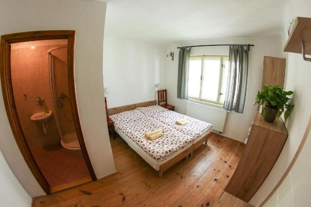 A bedroom with a wooden floor, a bed with a floral bedspread, a window with curtains, a plant on a wooden shelf, and a door leading to a bathroom.