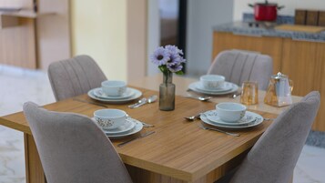Una mesa de comedor servida con tazas y platillos blancos, un florero con flores moradas y una cafetera en la encimera.