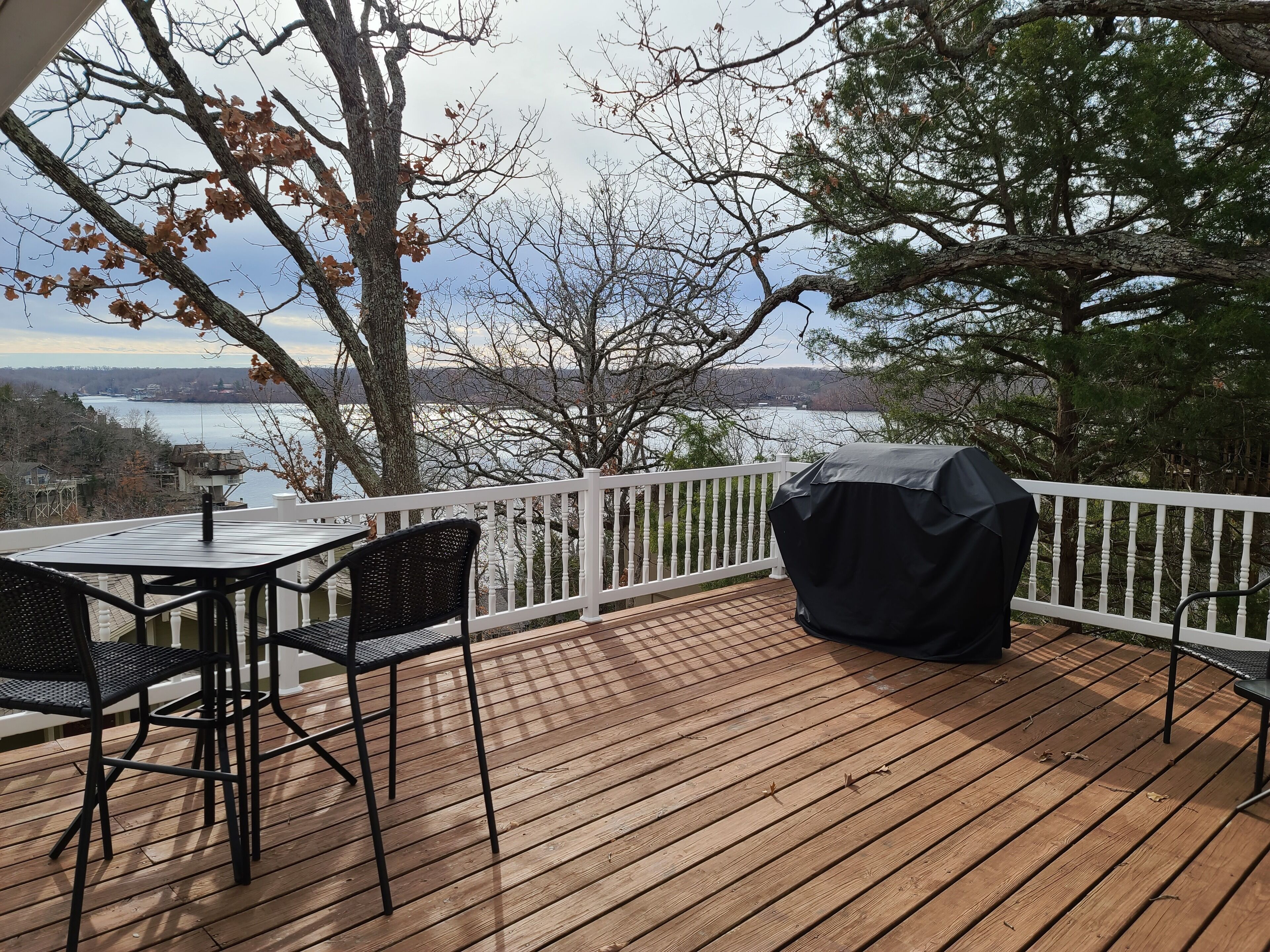 A wooden deck with a table and chairs, a barbecue grill, and a view of a lake and trees.