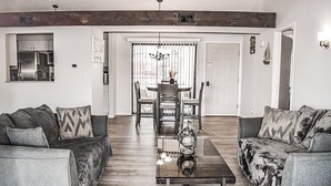 A modern living room with a glass coffee table, two grey sofas, and a dining area with a chandelier.