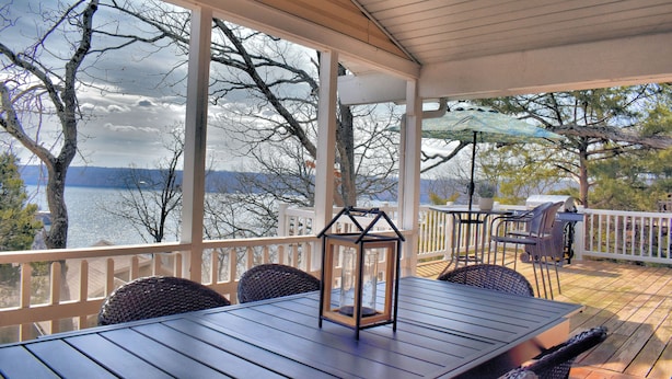 A covered outdoor deck with a table and chairs, overlooking a body of water and trees.