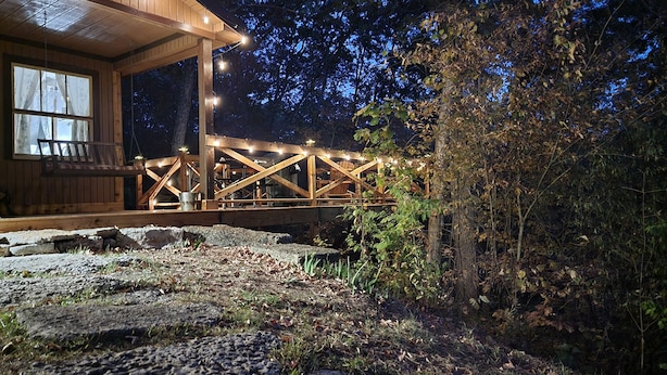 A wooden cabin with a porch and string lights, surrounded by trees and a stone pathway.