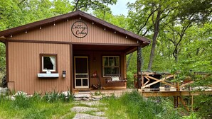 A rustic cabin labeled "Cottage Cabin" with a wooden exterior, a porch with a bench, and a stone pathway leading to the entrance.