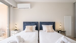 Hotelkamer met twee bedden, witte lakens en bijpassende hoofdeinden. Boven de bedden hangt een airconditioner, en aan beide kanten staat een nachtkastje met een lamp.