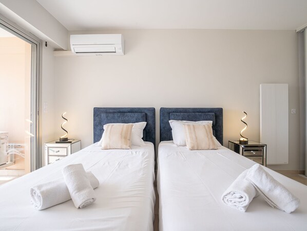 Hotelkamer met twee bedden, witte lakens en bijpassende hoofdeinden. Boven de bedden hangt een airconditioner, en aan beide kanten staat een nachtkastje met een lamp.