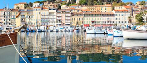 A marina with boats docked, a wooden pier, and a backdrop of colorful buildings and a hill with a tower.