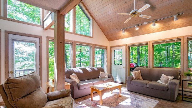 Ein geräumiges Wohnzimmer mit zwei Sofas, einem Couchtisch und einem Deckenventilator. Große Fenster bieten natürliches Licht und Blick auf den umliegenden Wald.