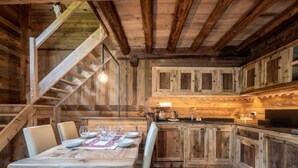Un interior rústic de fusta amb una taula de dinar per a quatre, una zona de cuina amb armaris de fusta, i una escala que condueix a un nivell superior.