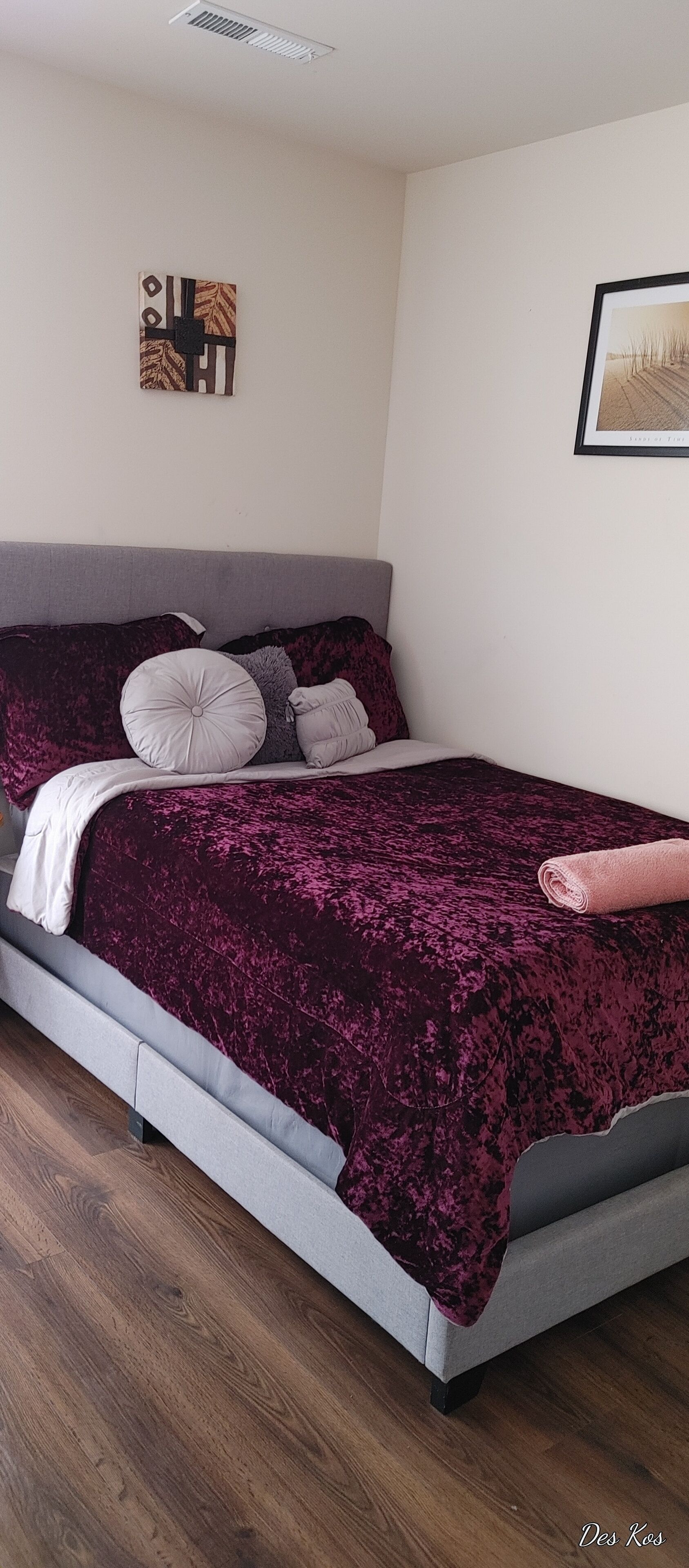 Een moderne slaapkamer met een grijs hoofdeinde, een paars fluweelbedovertrek en een keurig opgevouwen roze handdoek op het bed.