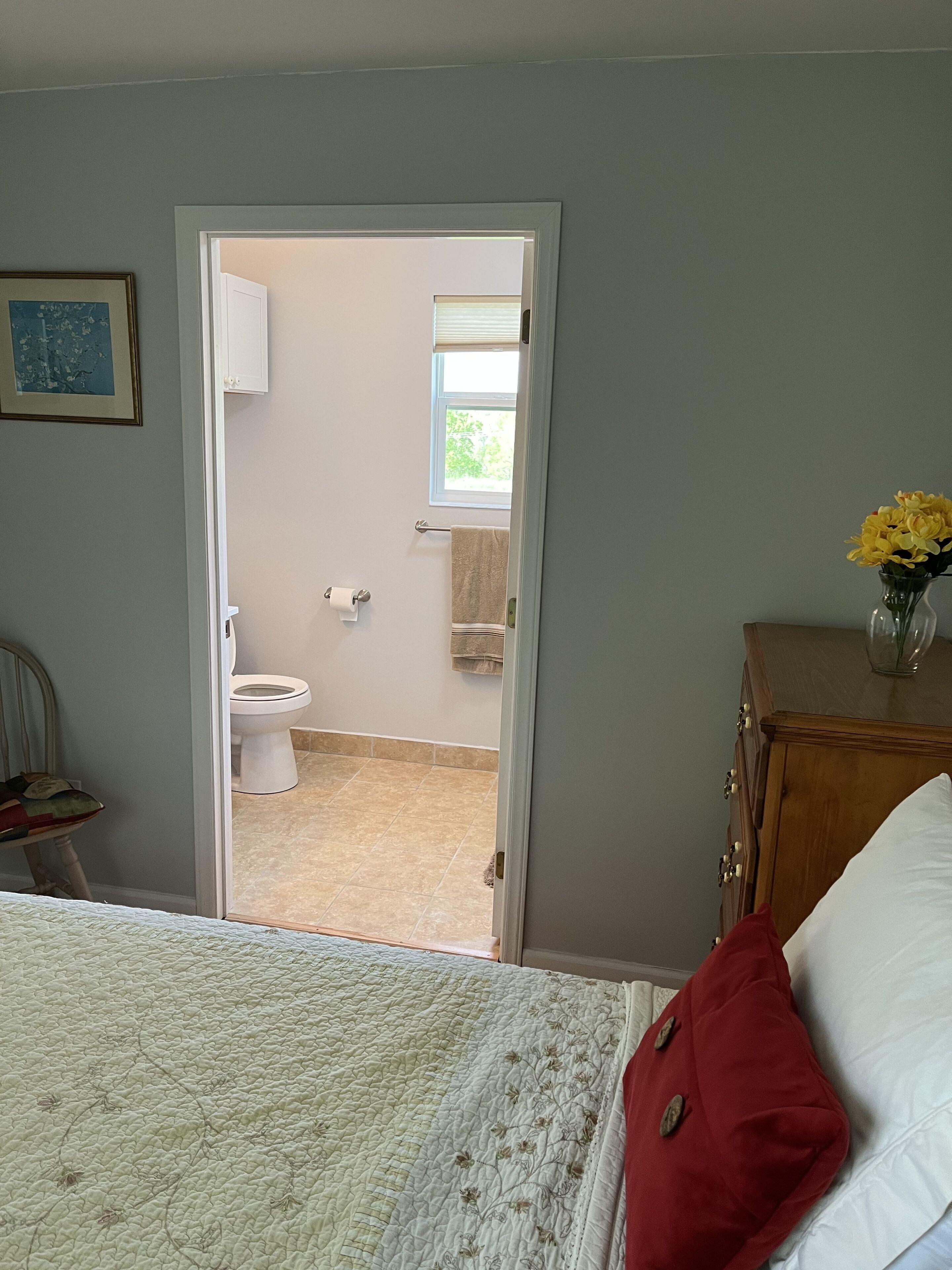 Un dormitorio con cama, cómoda de madera, un jarrón con flores y una puerta que da a un baño con inodoro y ventana.