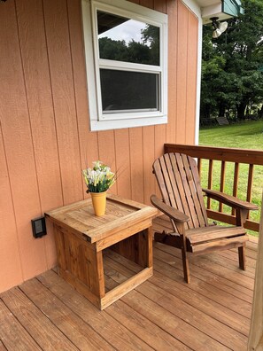 Um deck de madeira com uma pequena mesa e cadeira, uma janela e um vaso de planta.