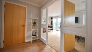 Un pasillo con una puerta de madera, un estante blanco con una planta, y vista a una habitación con un escritorio y una silla.
