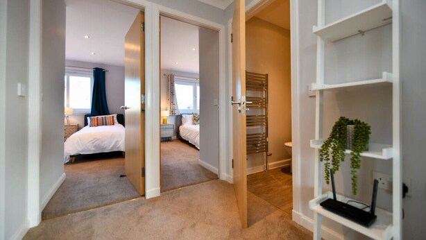 Habitación de hotel con cama, mesita de noche, cortinas en la ventana, radiador y estante blanco con una planta.