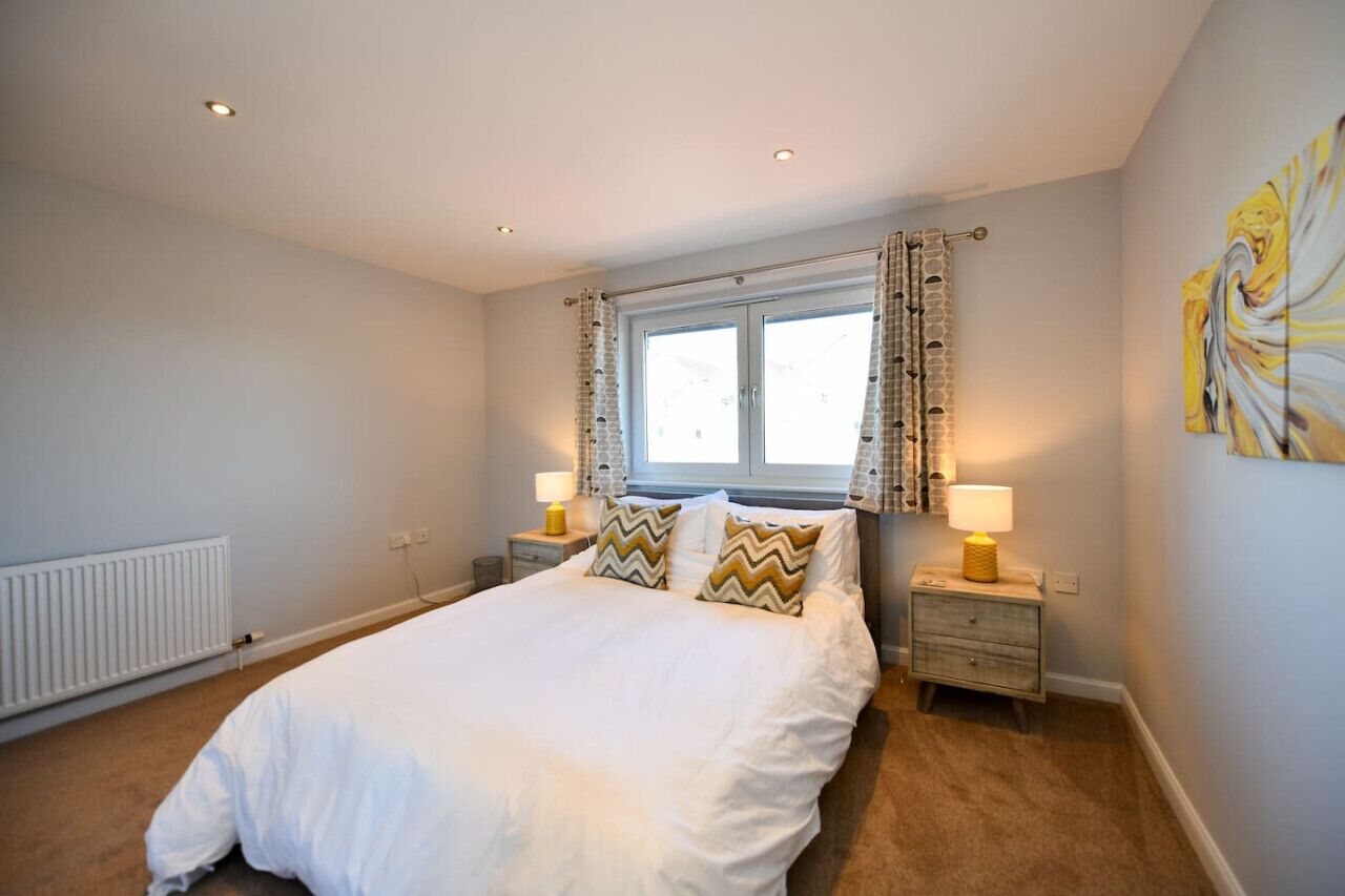 A bedroom with a large bed, two bedside tables with lamps, a window with curtains, a radiator, and a painting on the wall.