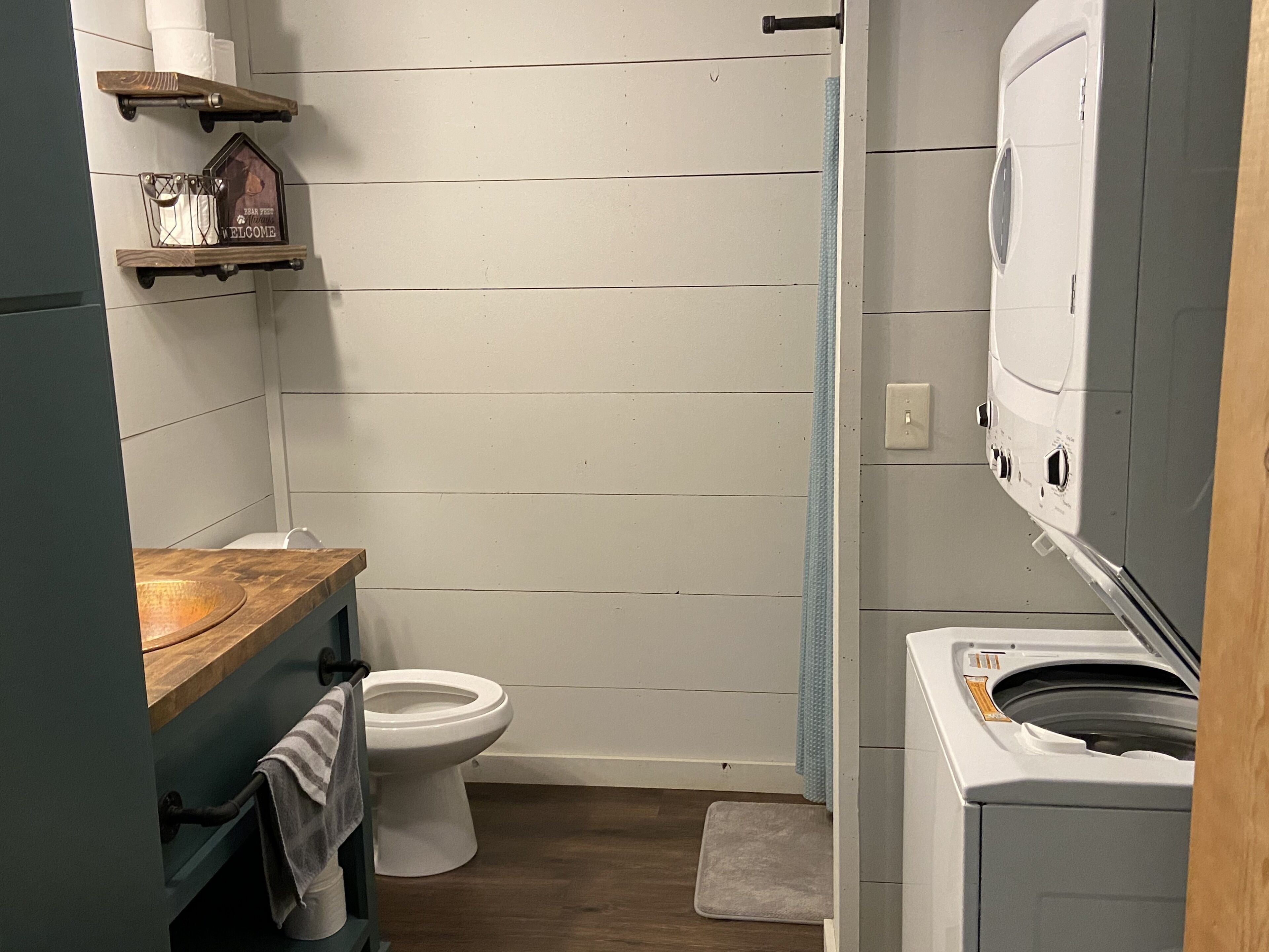 Een compacte badkamer met een houten wastafelmeubel, toilet, wasmachine en een klein plankje met een decoratief object.