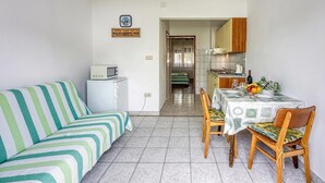 Een knus woongedeelte met een gestreept bankje, een eettafel met stoelen, een klein keukentje en een deur die naar een andere kamer leidt.