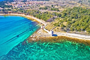 An aerial view of a coastal area with clear turquoise waters, a stone structure, and a village in the background.
