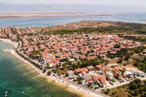 An aerial view of a coastal town with red-roofed buildings, a beach, and a clear view of the sea and mountains in the background.