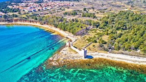 An aerial view of a coastal area with clear turquoise waters, a stone structure, and a village in the background.