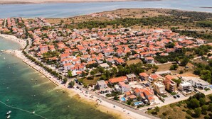 An aerial view of a coastal town with red-roofed buildings, a beach, and a clear view of the sea and mountains in the background.
