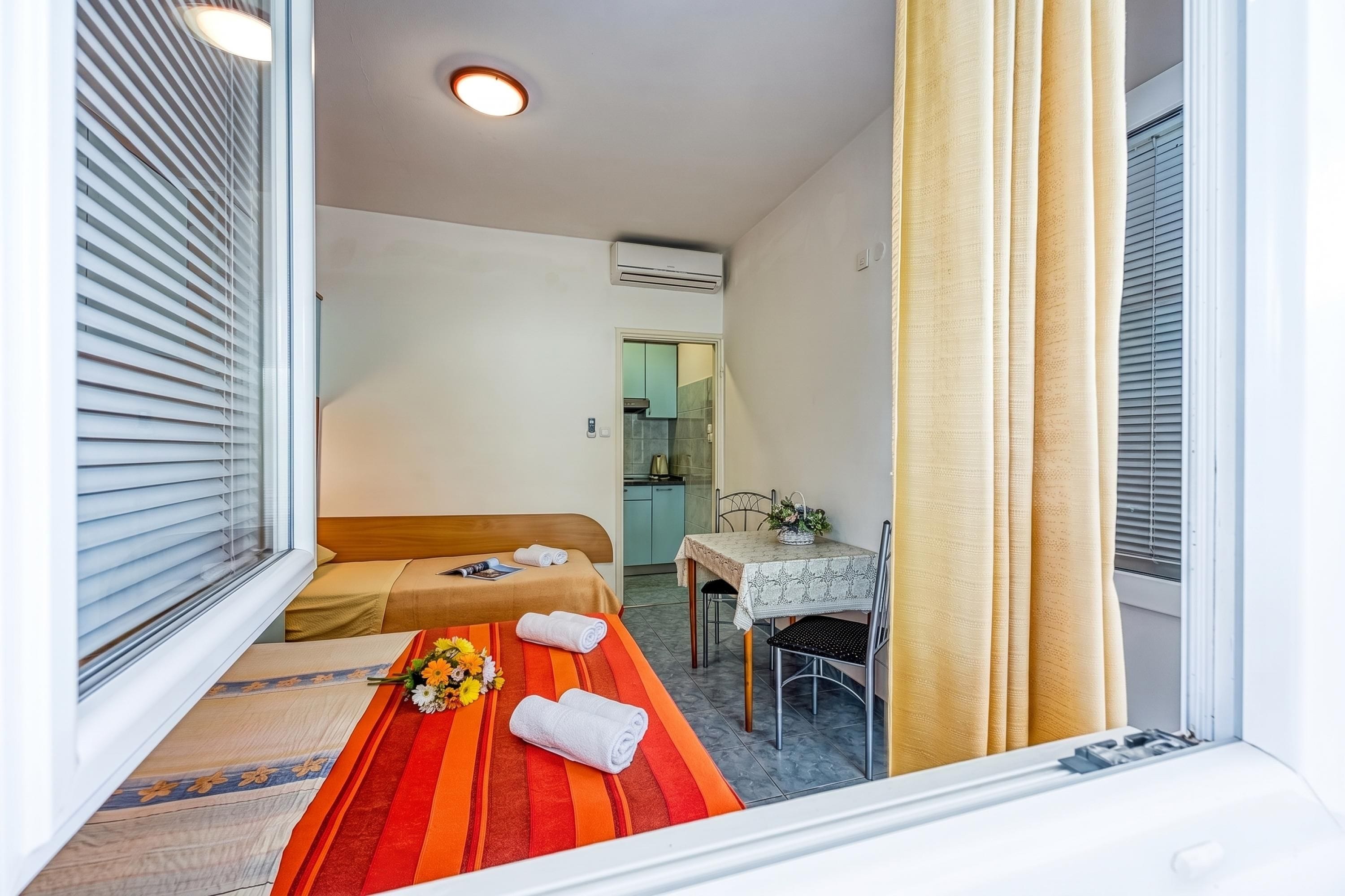 Une chambre d’hôtel avec un lit, une table recouverte d’une nappe, une chaise et une fenêtre avec des stores.