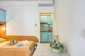 A small, clean room with a bed, a table with a floral arrangement, and a kitchenette visible through an open door.