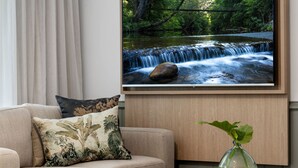 Un salón con un sofá beige, un televisor de pantalla plana grande montado en la pared que muestra una cascada, y una mesita redonda con un jarrón transparente que contiene una planta.