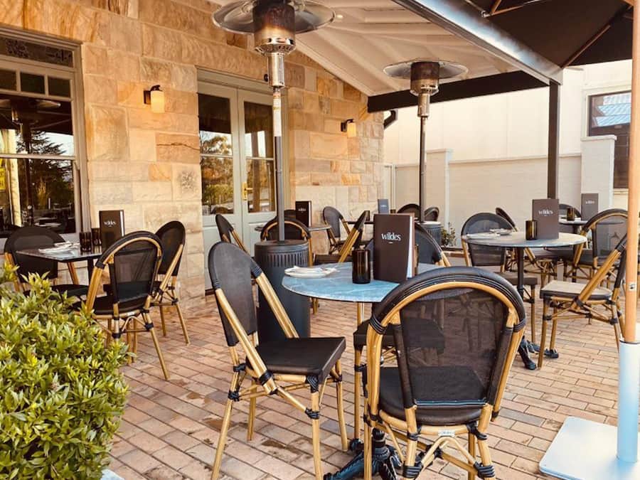 An outdoor patio with wicker chairs and tables, stone walls, and a menu stand.
