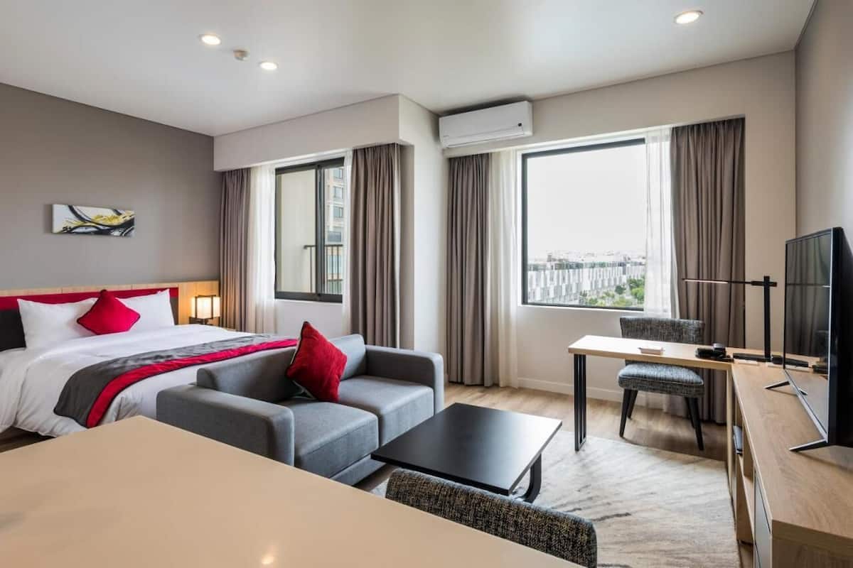 Une chambre d’hôtel moderne avec un lit, un canapé gris, un fauteuil, une petite table et un téléviseur.