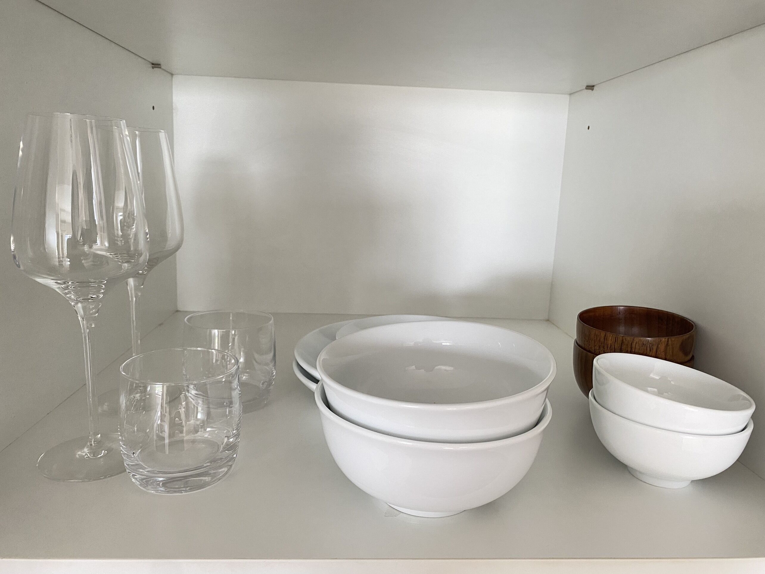 A white shelf with clear wine glasses, white bowls, and a wooden bowl.