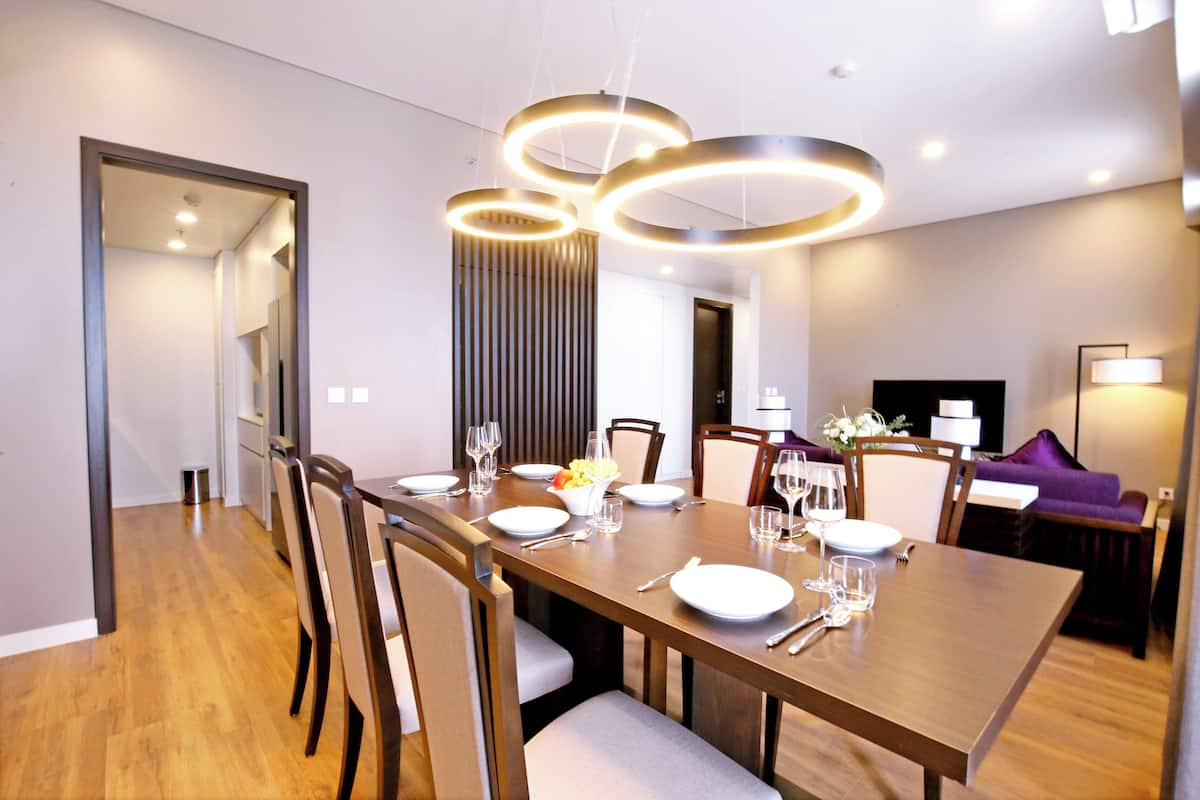 Un espace de salle à manger moderne, avec une longue table en bois dressée pour six personnes, entourée de chaises. Des luminaires suspendus sont placés au-dessus de la table, et on aperçoit un téléviseur à l’arrière-plan.