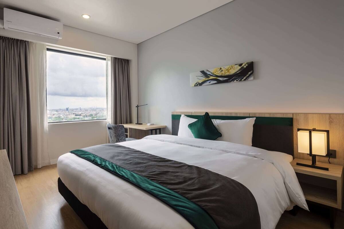 Une chambre d’hôtel avec un grand lit, un bureau, une chaise, une table de chevet avec une lampe, un tableau au mur et une vue sur la ville par la fenêtre.