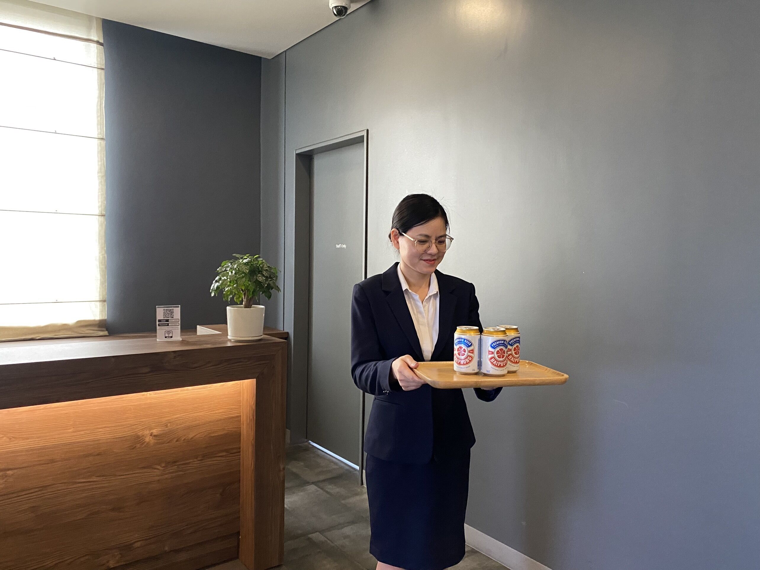 A person holding a tray with two cans of beer in a modern office setting with a wooden reception desk and a potted plant.