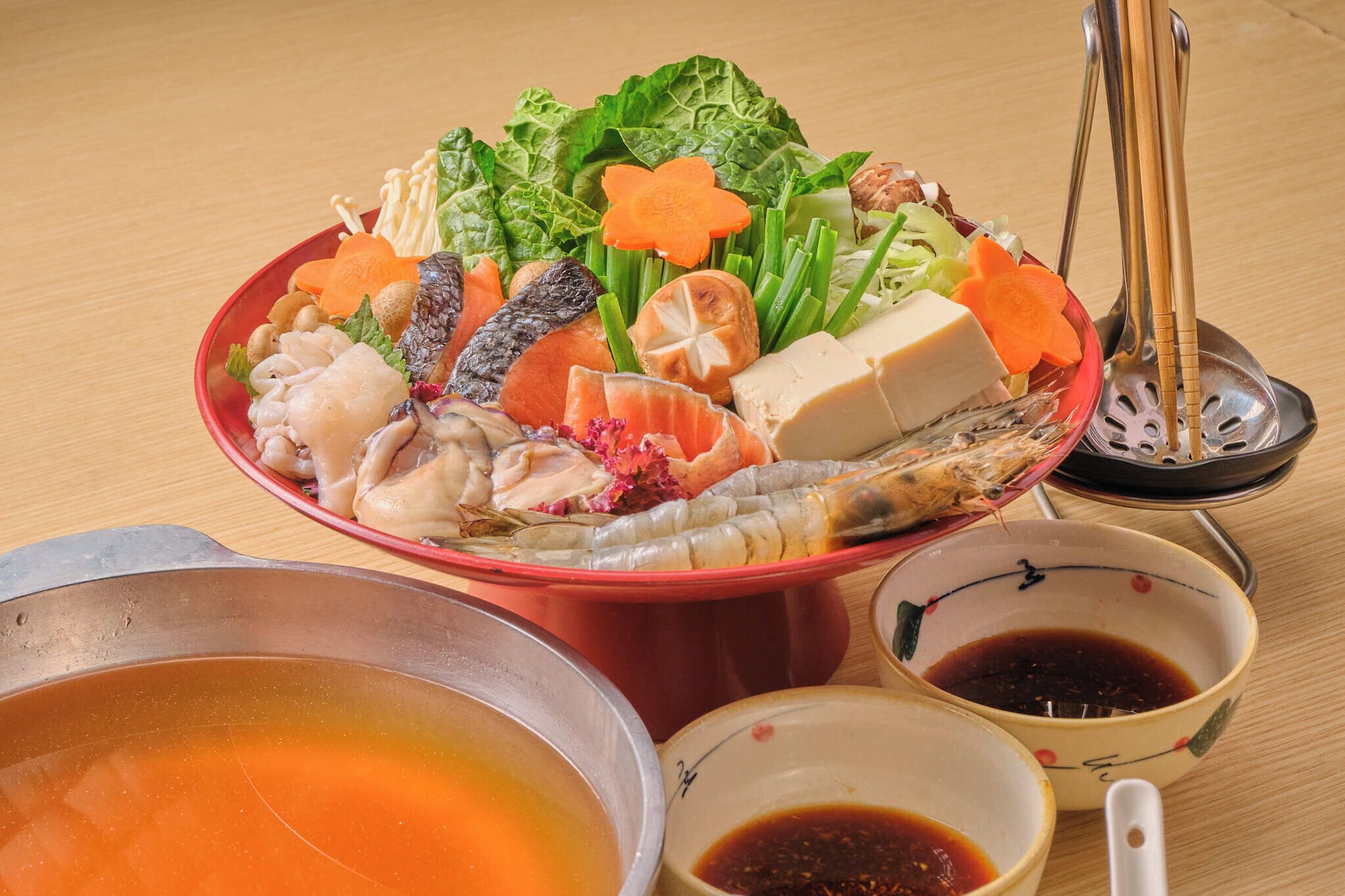 A bowl of assorted seafood and vegetables with dipping sauces on a wooden table.