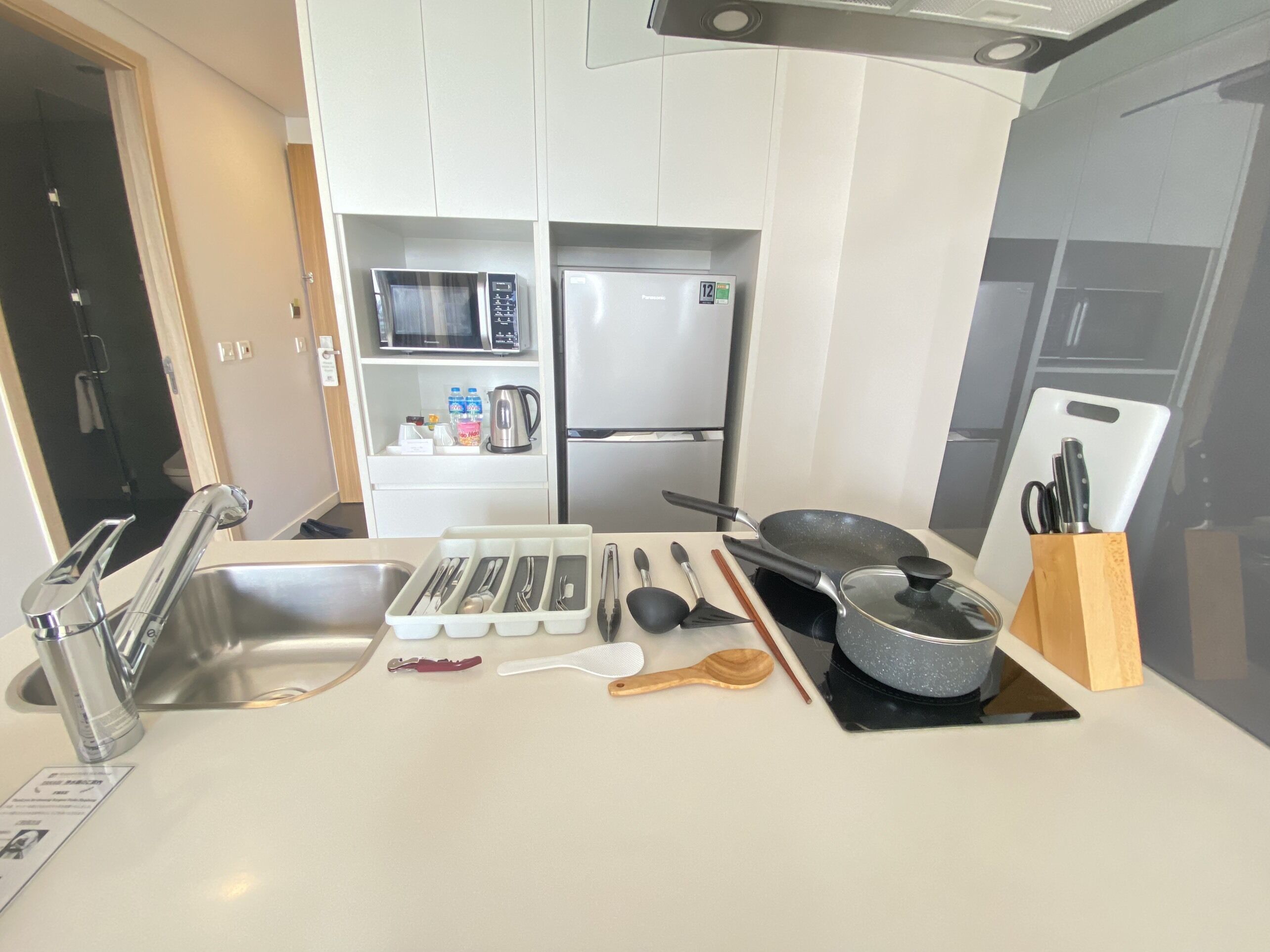 Una cocina moderna con armarios blancos, un lavamanos de acero inoxidable, un microondas, un refrigerador y diversos utensilios de cocina en la encimera.