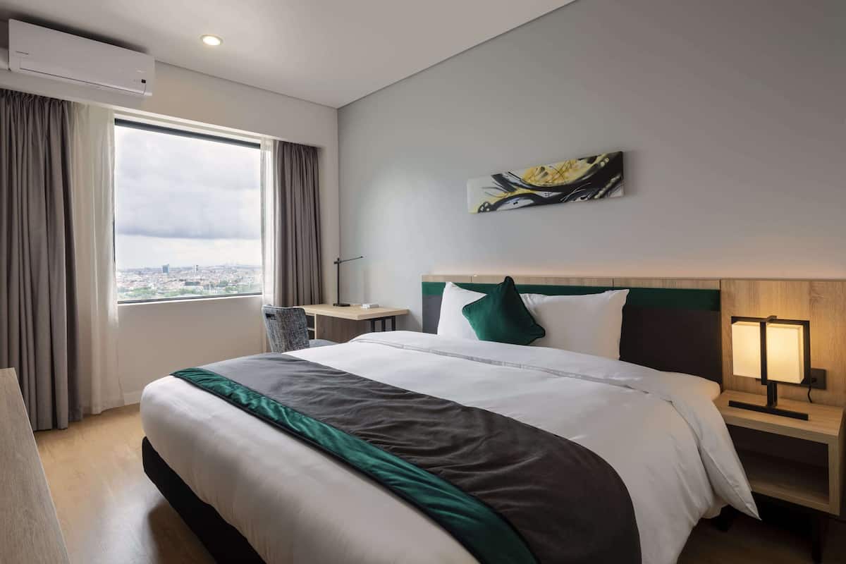Une chambre d’hôtel avec un grand lit, une table de chevet avec une lampe, une fenêtre avec des rideaux et une vue sur la ville.